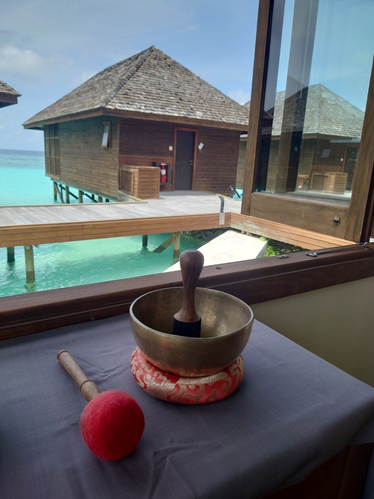 A serene view featuring a wooden building on stilts over turquoise water, with a singing bowl and a red mallet placed on a table in the foreground.